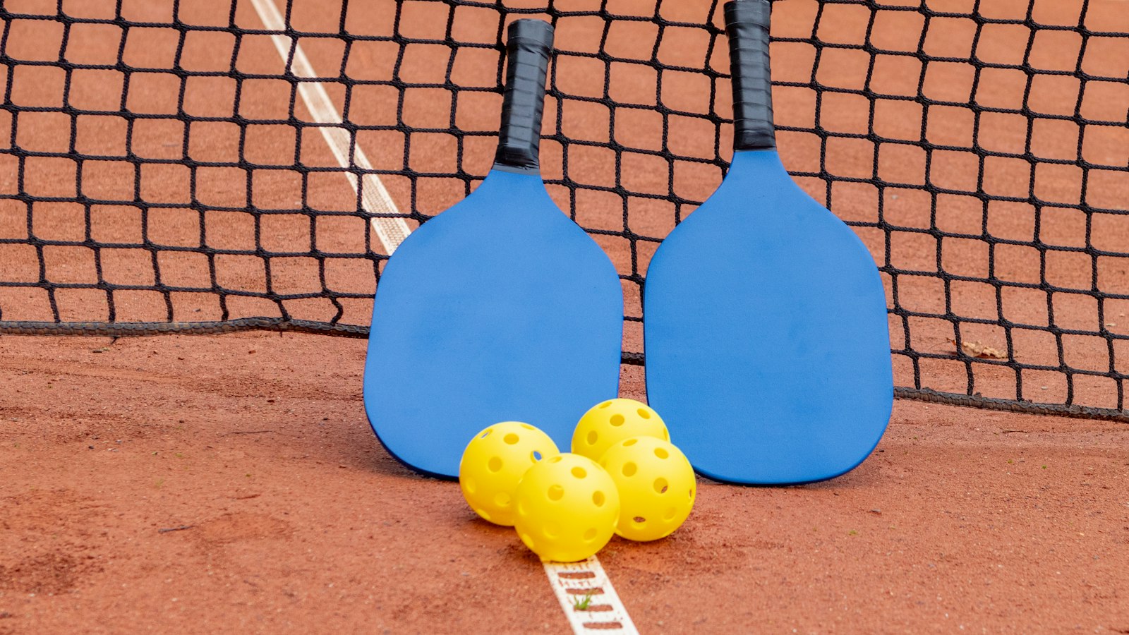Two pickleball paddles and three balls resting against the net on a court